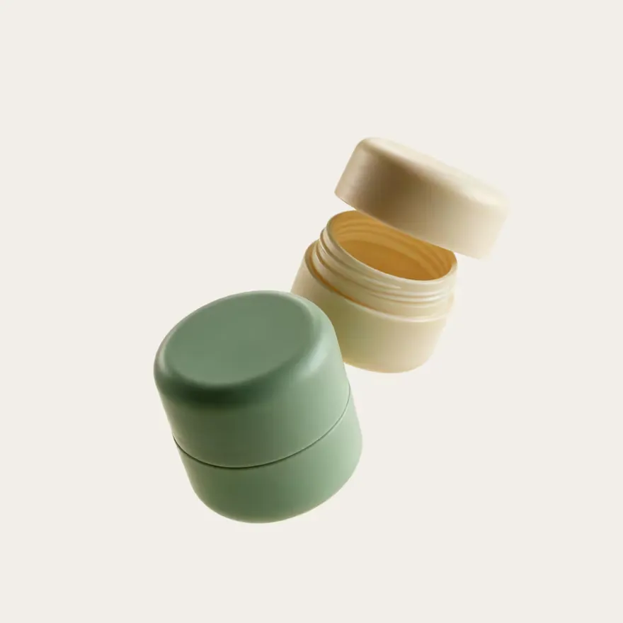 A closed green ointment jar beside an open cream-coloured one.