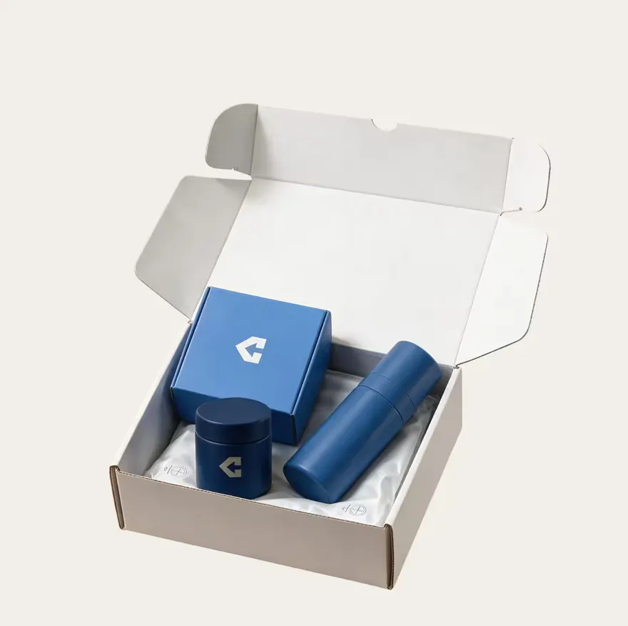 An open corrugated mailer packed with a branded box, a jar and a bottle on a foil liner.